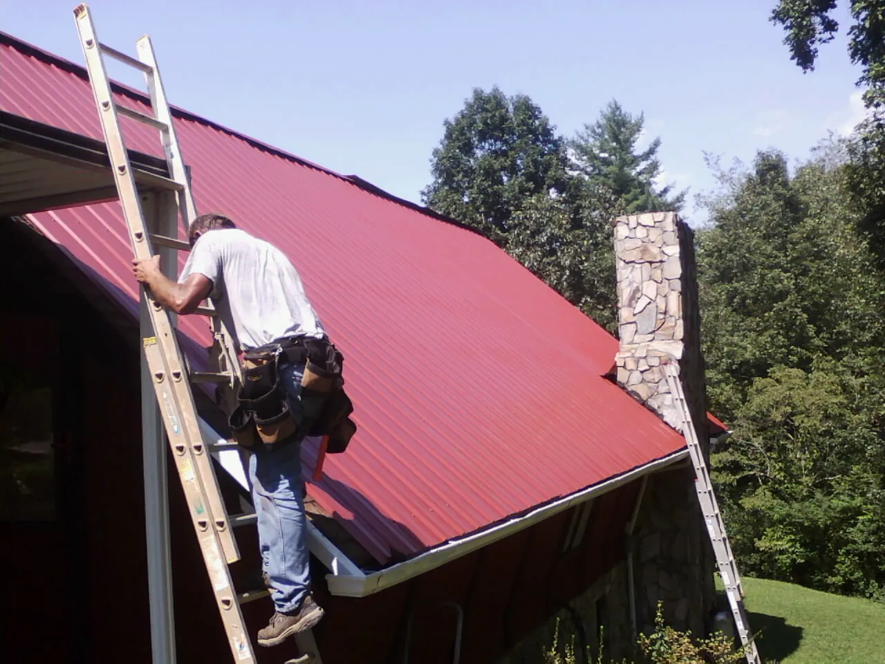 Roofer on a ladder beside a red metal roof and stone chimney — Soffit & Fascia Repair by Roofing Brothers in Sweet Springs